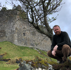 Scottish Gaelic consultant Àdhamh Ó Broin kneeling in front of a castle in the Scottish Highlands
